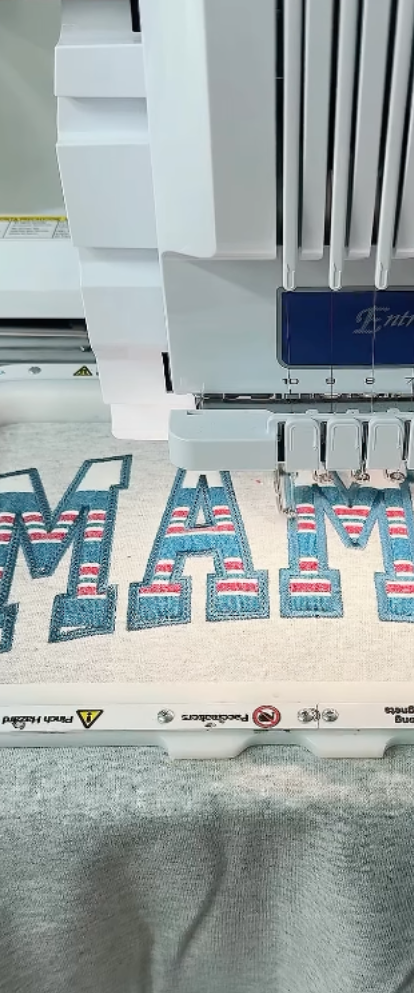Embroidery machine sewing 'MAMA' onto the sweater with baby clothes fabric.